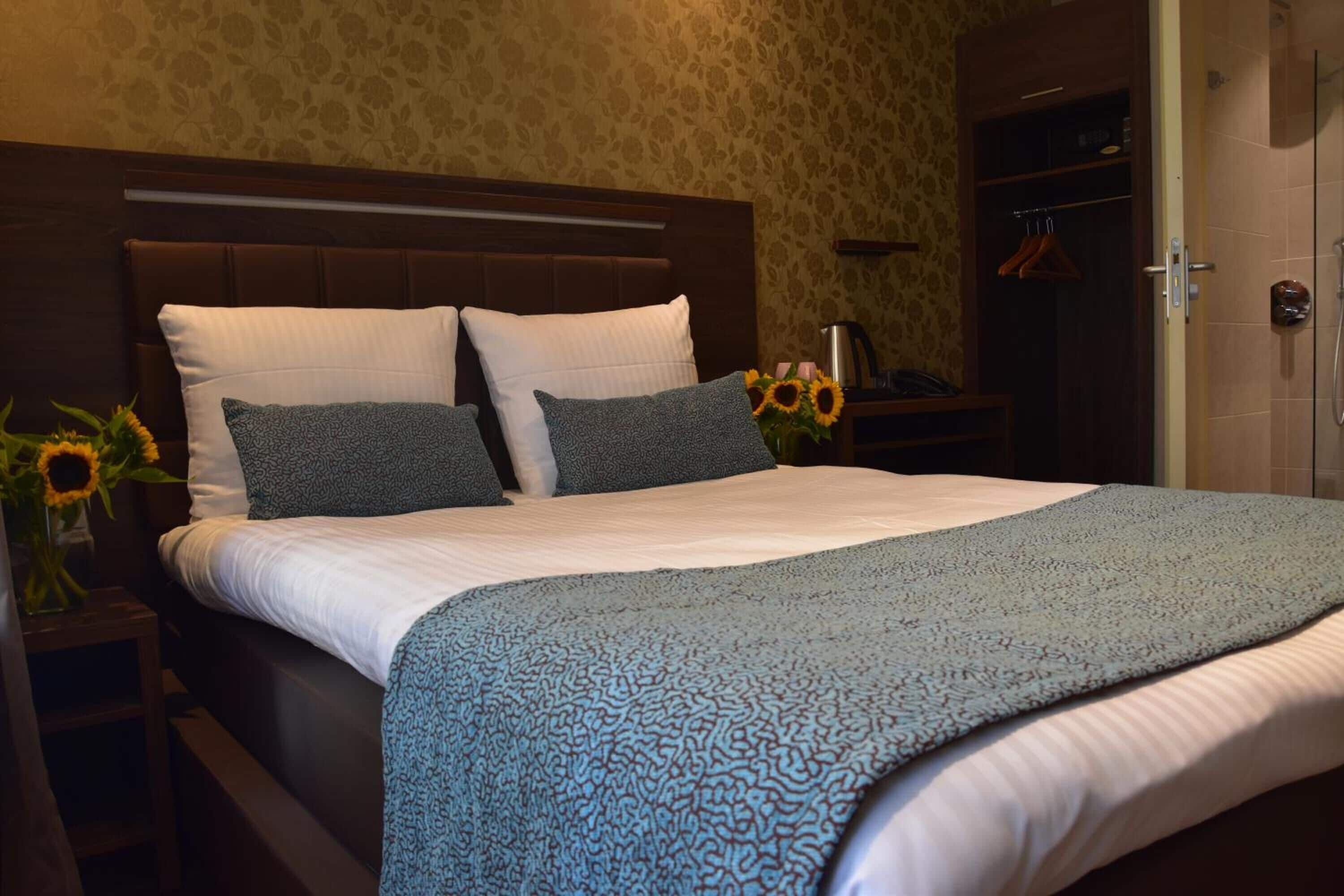 Een netjes opgemaakt bed met een bedsprei en kussens met een patroon, een nachtkastje met een vaas zonnebloemen, en een houten hoofdbord.