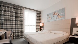 A hotel room with two beds, a desk, and a chair. There is a painting on the wall and a lamp on the nightstand.