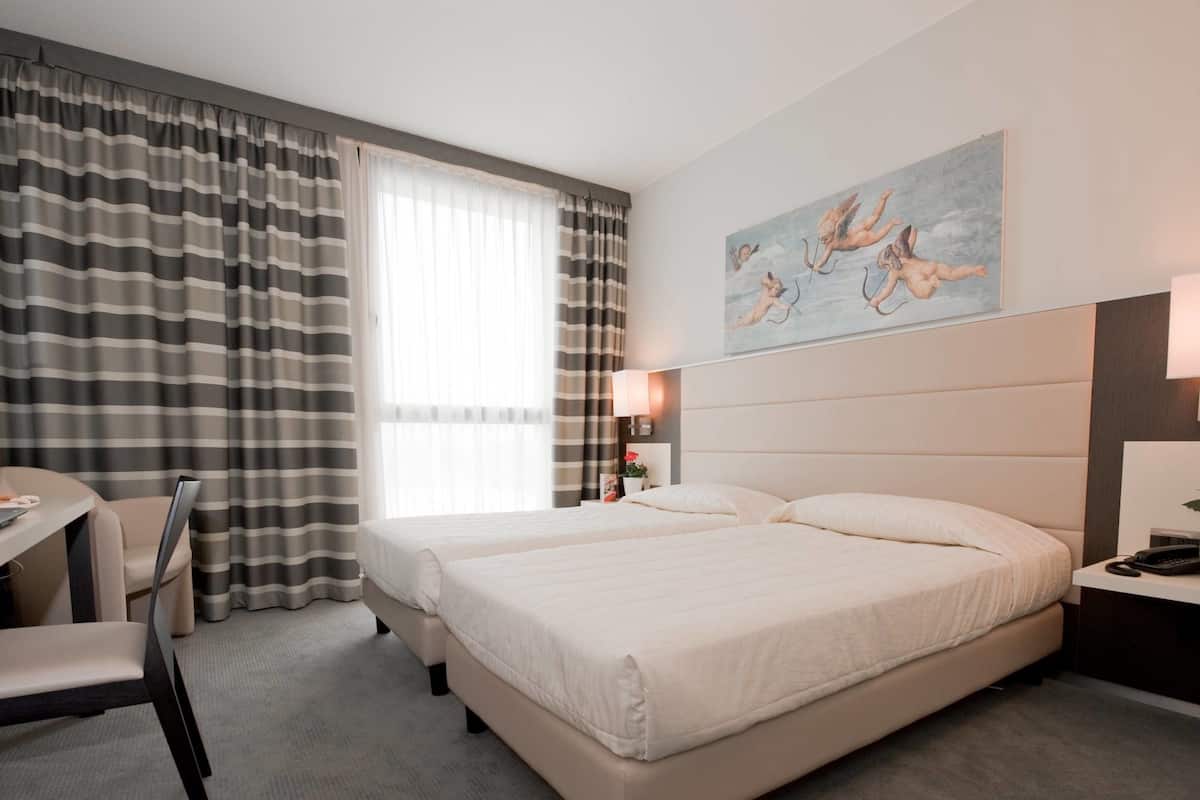 Camera d'albergo con due letti, una scrivania e una sedia. Sul muro c'è un quadro e un comodino con una lampada.