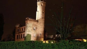 Una construcción al estilo de castillo con una torre destacada, rodeada de vegetación e iluminada con luces artificiales.
