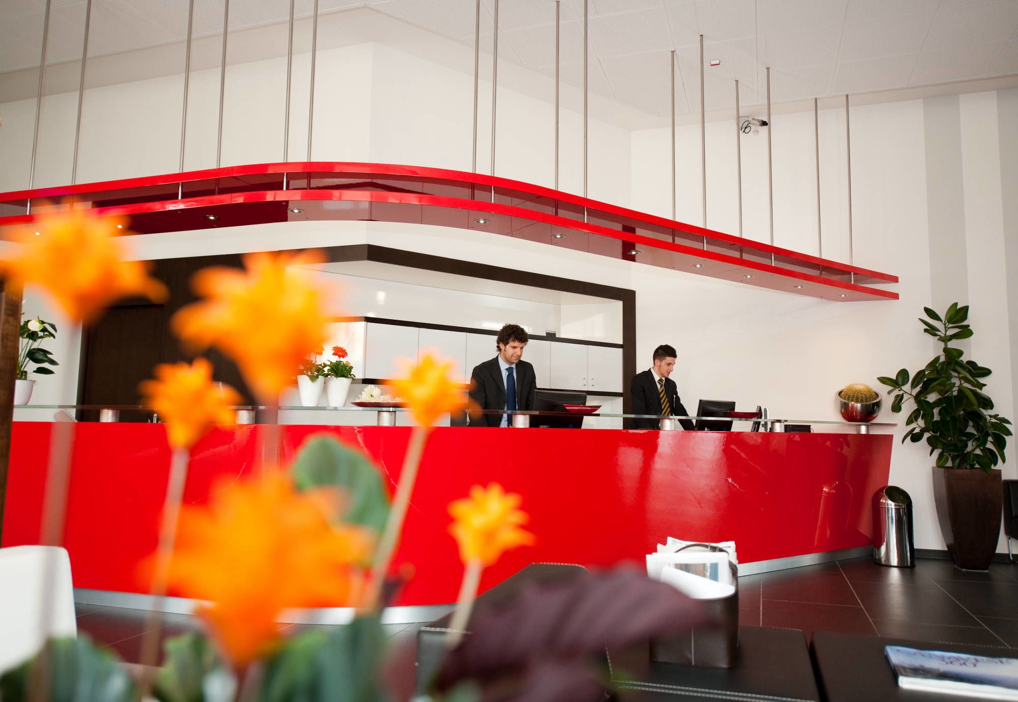 Un'area reception di un hotel con un bancone rosso, due addetti alla reception e un design d'interni moderno.