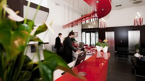 A modern hotel lobby with a red reception desk, a staircase, and a seating area with plants.