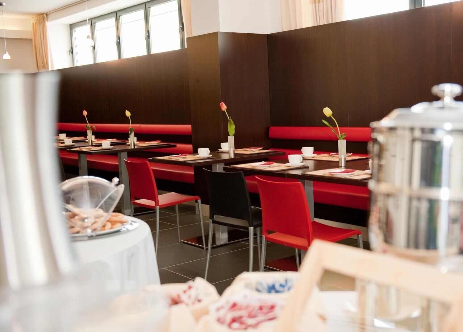 Ein Restaurant mit roten Sitzbänken und schwarzen Stühlen, Tische mit weißen Tassen und Untertassen sowie eine Kaffeekanne auf dem Tisch.