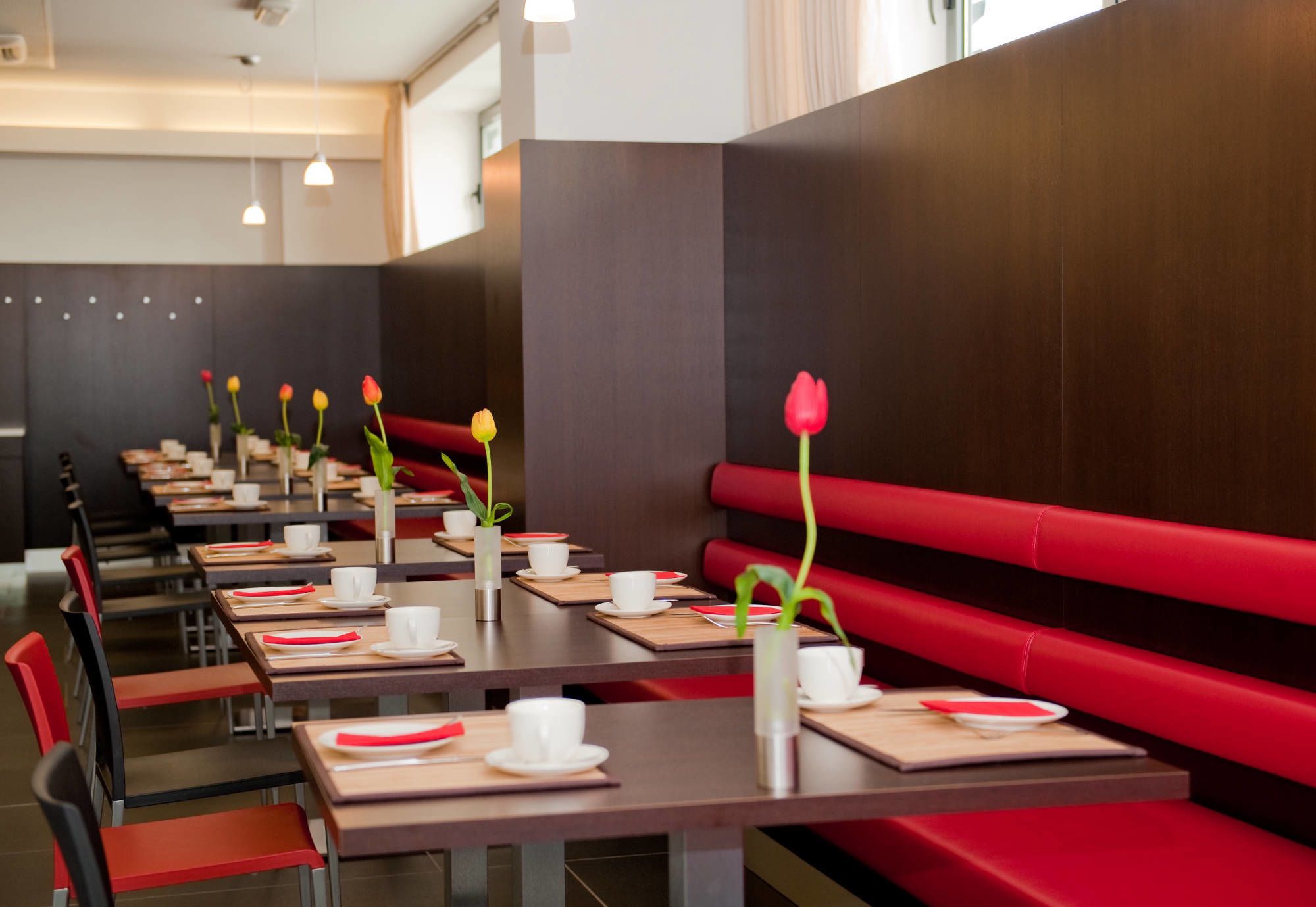 Un ristorante con panche rosse e lunghi tavoli, ognuno apparecchiato con tazze e piatti bianchi, e vasi con fiori.
