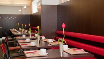 Un ristorante con panche rosse e lunghi tavoli, ognuno apparecchiato con tazze e piatti bianchi, e vasi con fiori.