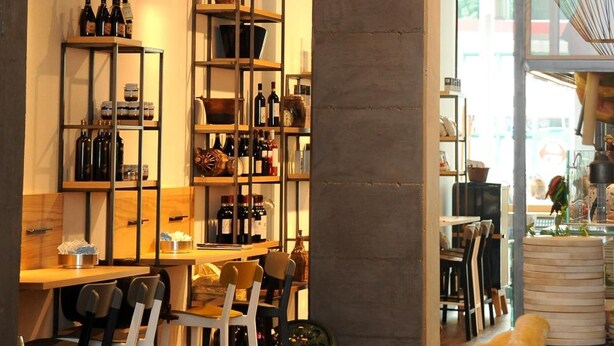 A restaurant interior with wooden tables and chairs, shelves with bottles, and a hanging light fixture.
