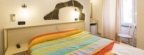 Habitación de hotel con cama, mesita de noche, termostato, luz en la pared, ventana con cortinas y una silla.