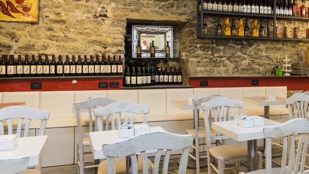 Un restaurant aux murs de pierre, avec des chaises et des tables blanches. On y trouve des bouteilles de vin sur des étagères et un miroir au mur.