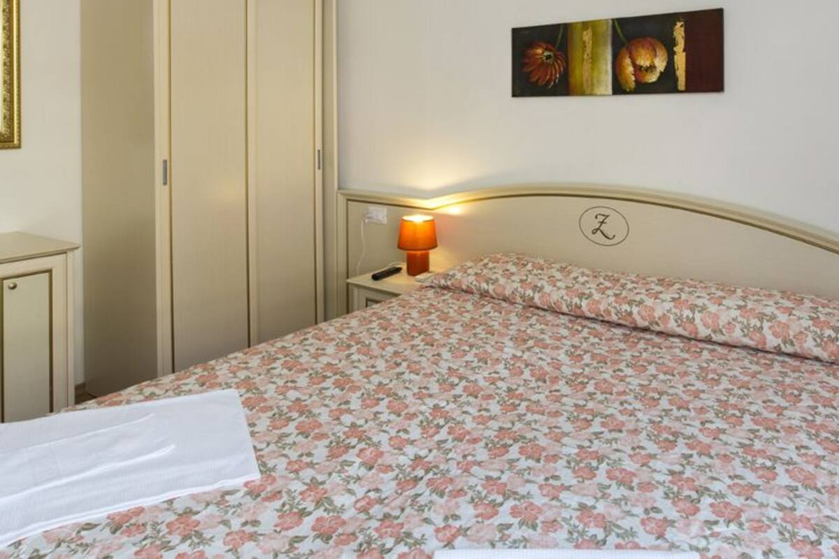 A neatly made bed with a floral bedspread, a bedside table with a lamp, a desk with a chair, and a mirror on the wall.