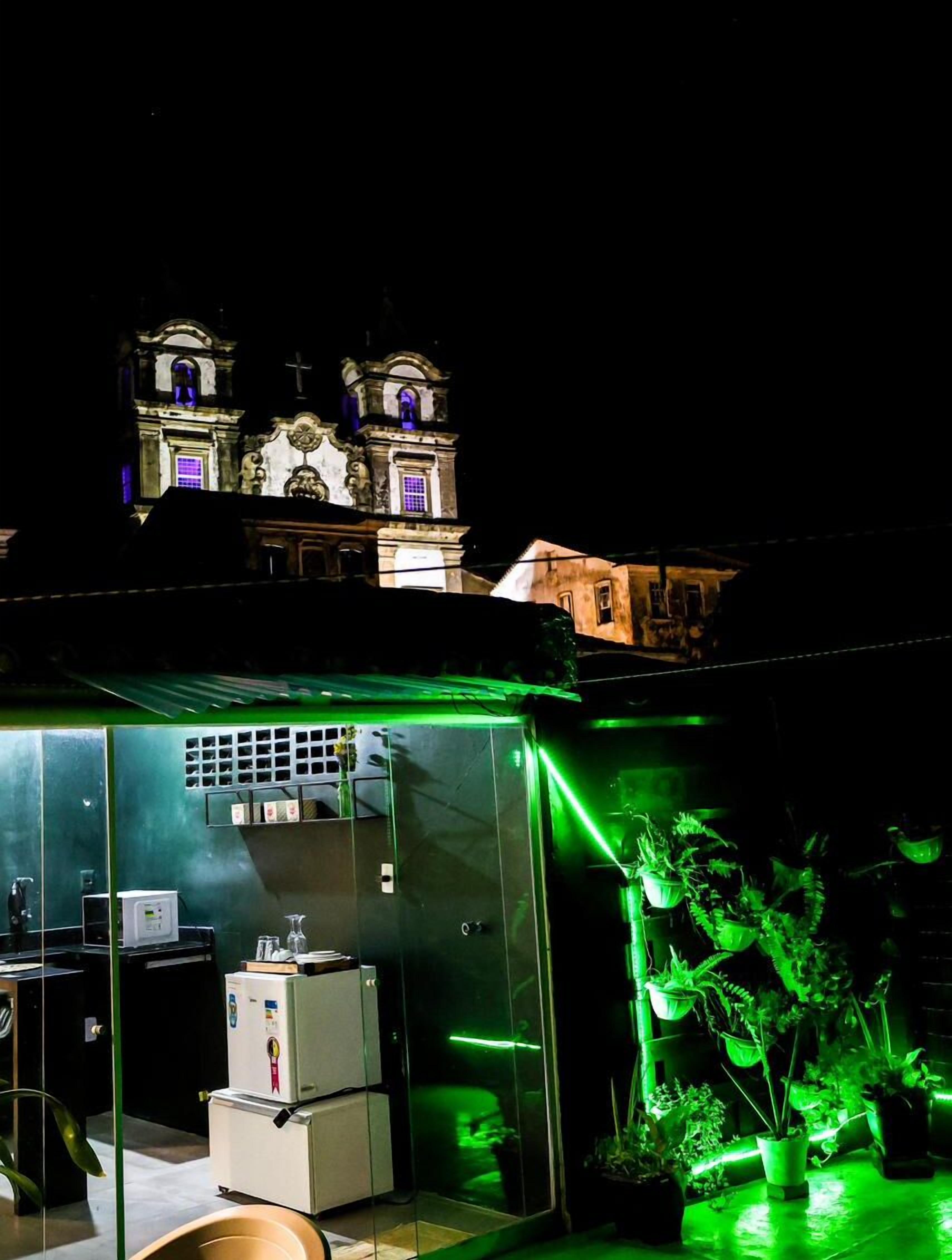 A building with a clock tower at night, a glass-walled room with a refrigerator and a computer, and green lighting.