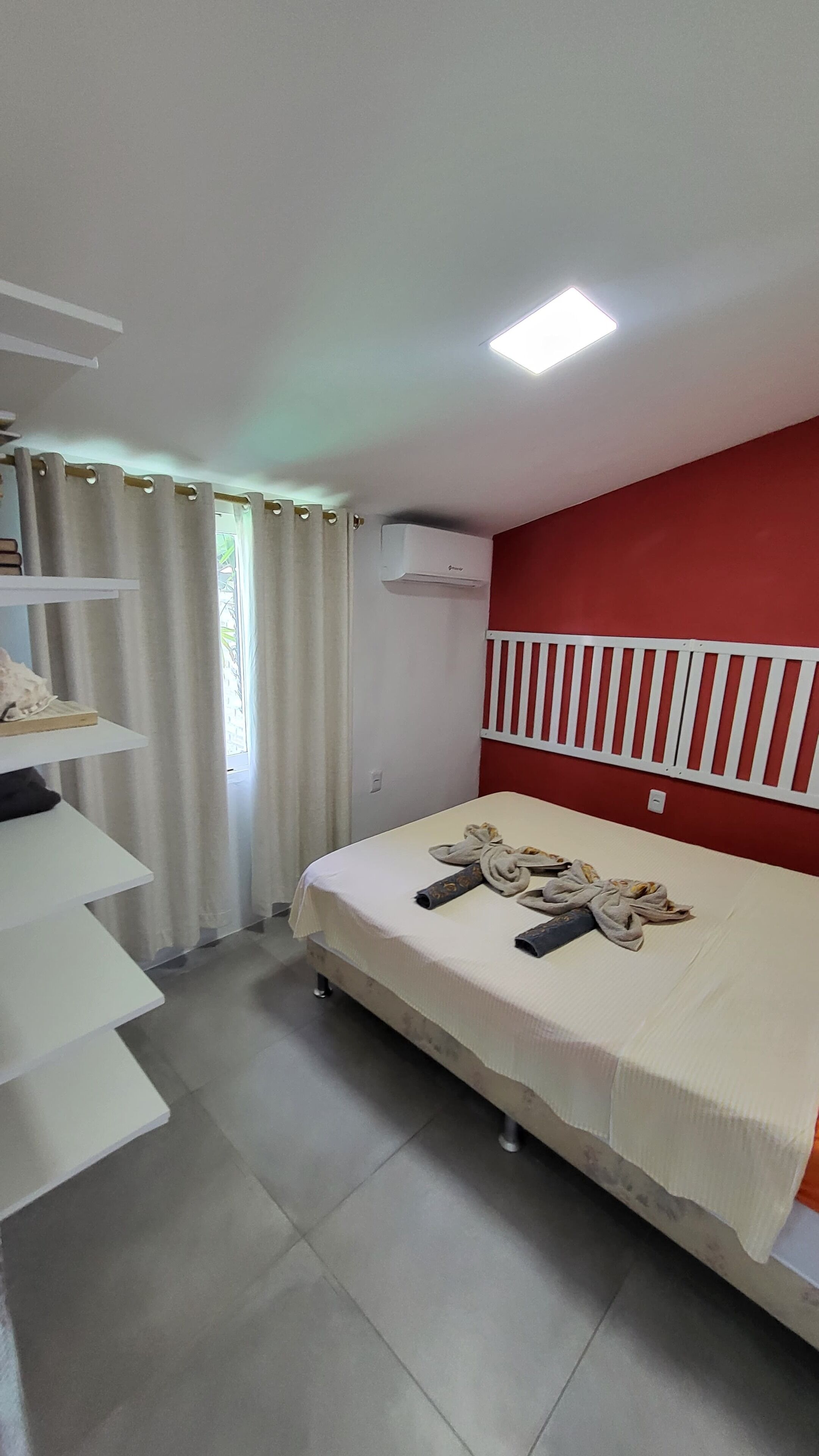 Een slaapkamer met een bed, een plank, een raam met gordijnen en een airconditioning.