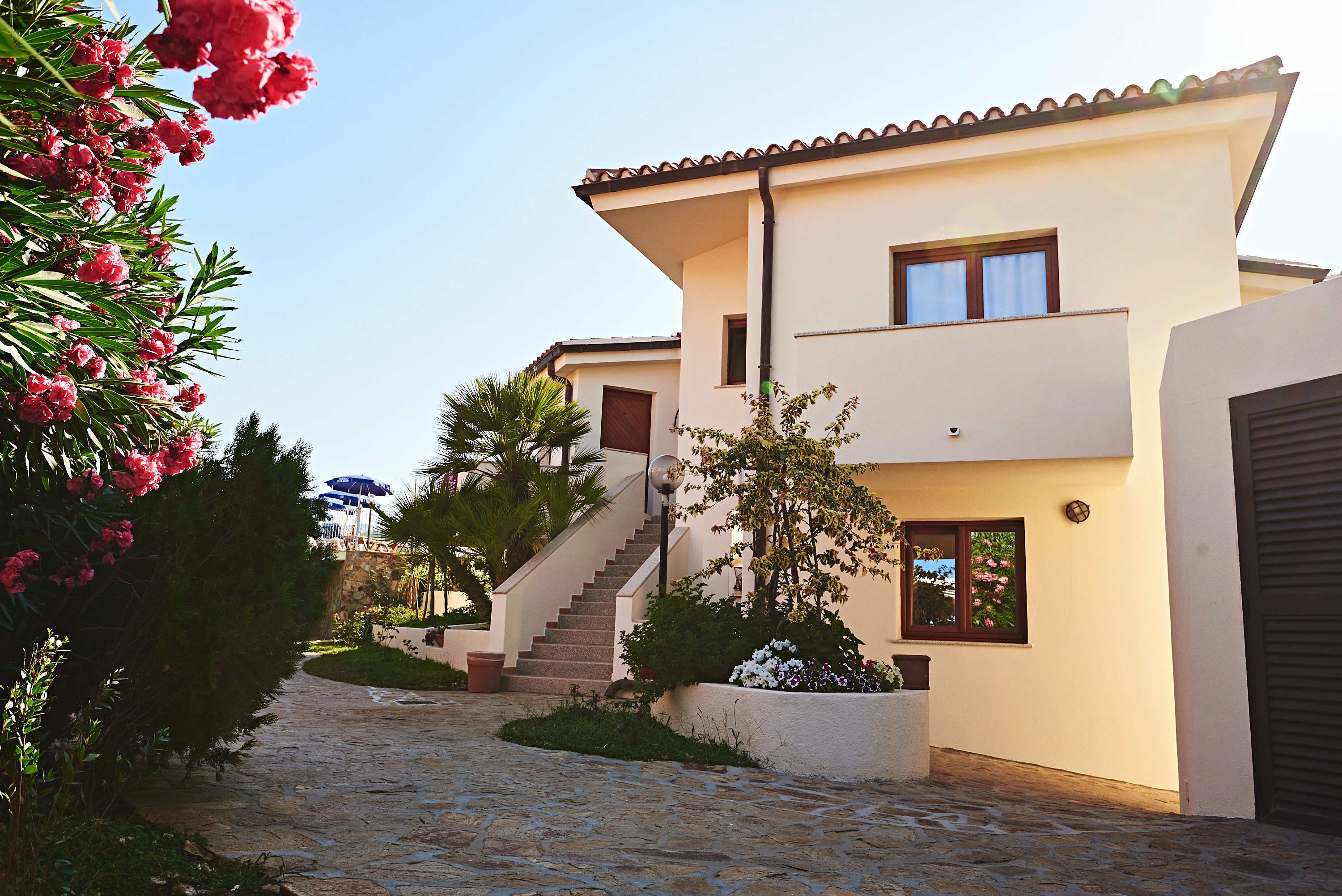 A modern two-story house with a stone pathway leading to the entrance, surrounded by lush greenery and flowering plants.