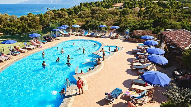 A swimming pool with multiple lanes, surrounded by lounge chairs and umbrellas, with a view of the sea and hills in the background.