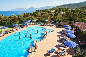 Una piscina con varias pistas de natación, rodeada de sillones de descanso y sombrillas, con vistas al mar y colinas al fondo.