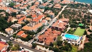 An aerial view of a coastal town with terracotta-roofed buildings, a marina, and a swimming pool.