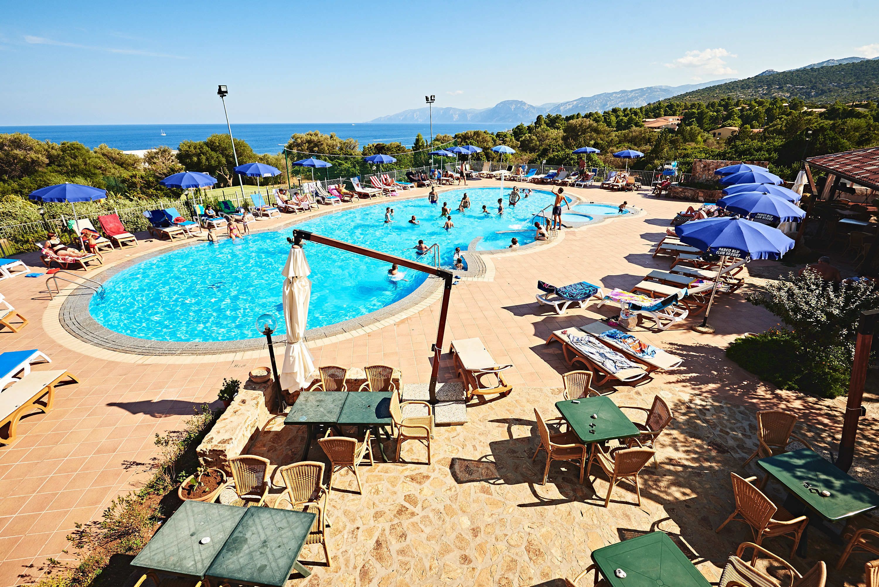 A pool area with lounge chairs and umbrellas, surrounded by a stone patio and a view of the sea.
