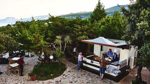 An outdoor dining area with stone flooring, wooden tables, and chairs, surrounded by lush greenery and a mountain view.