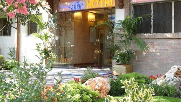 Entrada de hotel con fachada de vidrio, un letrero que dice "air HOTEL" y un jardín cuidado con plantas variadas y una roca grande.