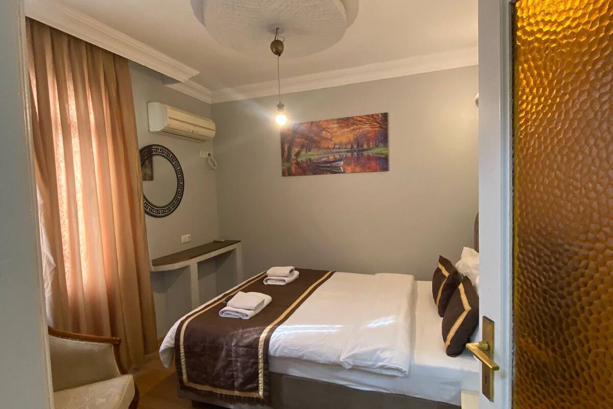 A hotel room with a bed, a bedside table, a chair, a painting on the wall, and a door with a textured glass panel.