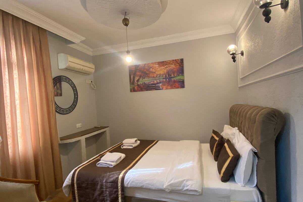 A hotel room with a bed, a desk, a chair, an air conditioning unit, a painting, and a ceiling light.