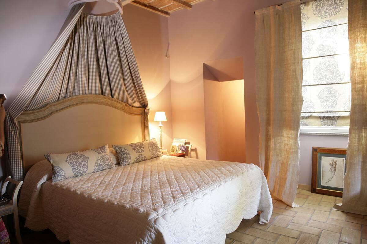 A neatly made bed with a canopy, a bedside table with a lamp, and a framed picture on the wall.