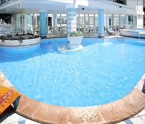 A large indoor swimming pool with clear blue water, surrounded by lounge chairs and a covered seating area. - Michelangelo (Cesenatico)