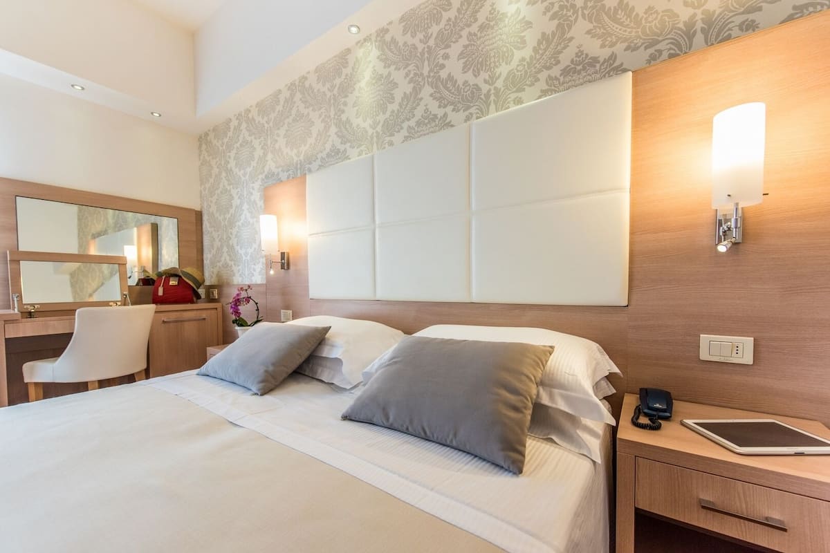 Habitación de hotel con una cama grande, un escritorio con silla, una mesita de noche con lámpara y una luz empotrada en la pared.