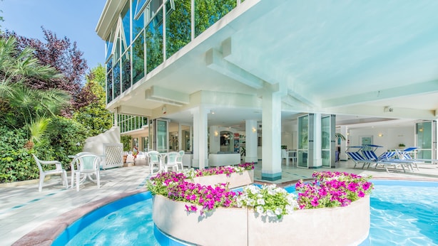 A swimming pool with a curved edge, surrounded by white lounge chairs and a covered area with glass doors. - Michelangelo (Cesenatico)