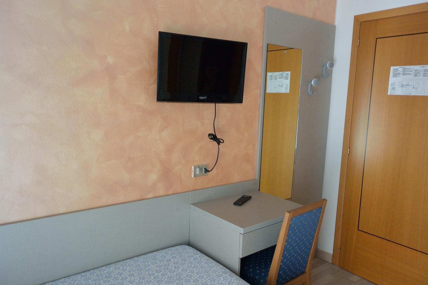 Quarto de hotel pequeno com uma cama, uma escrivaninha, uma cadeira, uma televisão fixada na parede e uma porta de madeira.