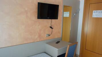 Quarto de hotel pequeno com uma cama, uma escrivaninha, uma cadeira, uma televisão fixada na parede e uma porta de madeira.