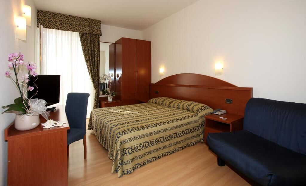 A hotel room with a bed, a desk with a chair, a sofa, a TV, and a mirror.