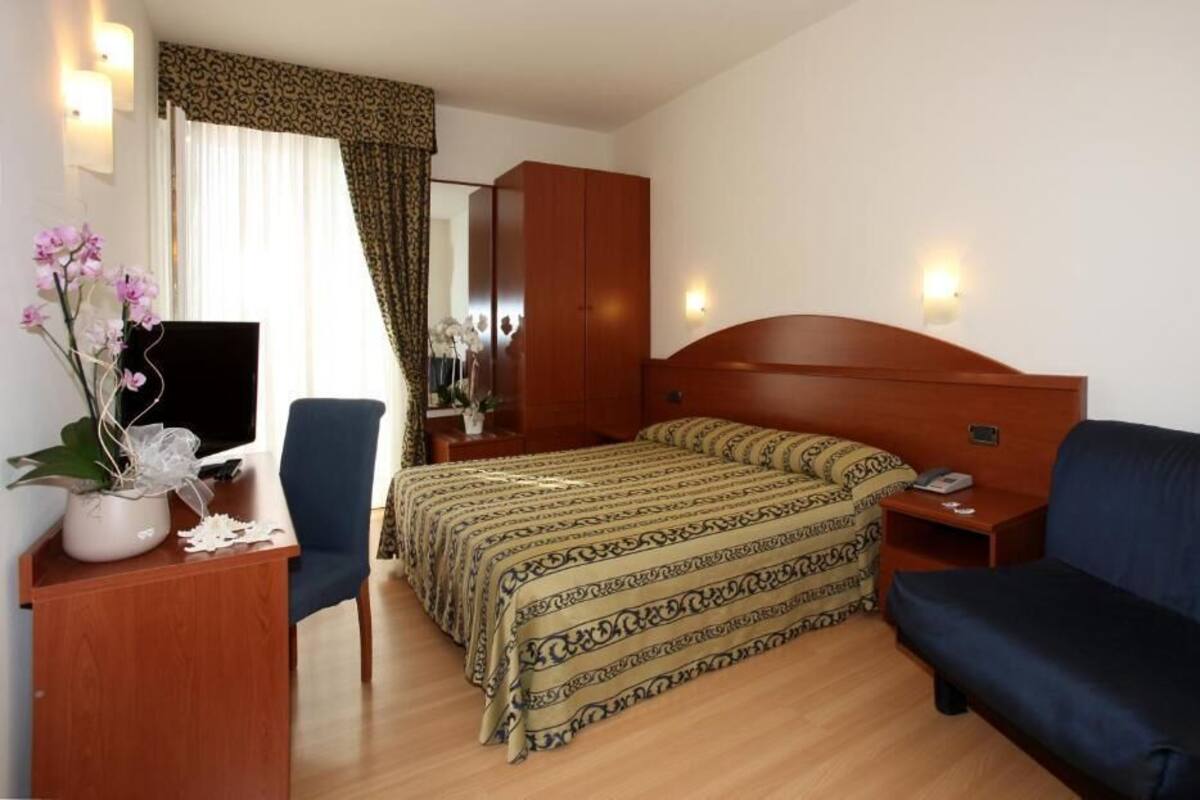 A hotel room with a bed, a desk with a chair, a sofa, a TV, and a mirror.