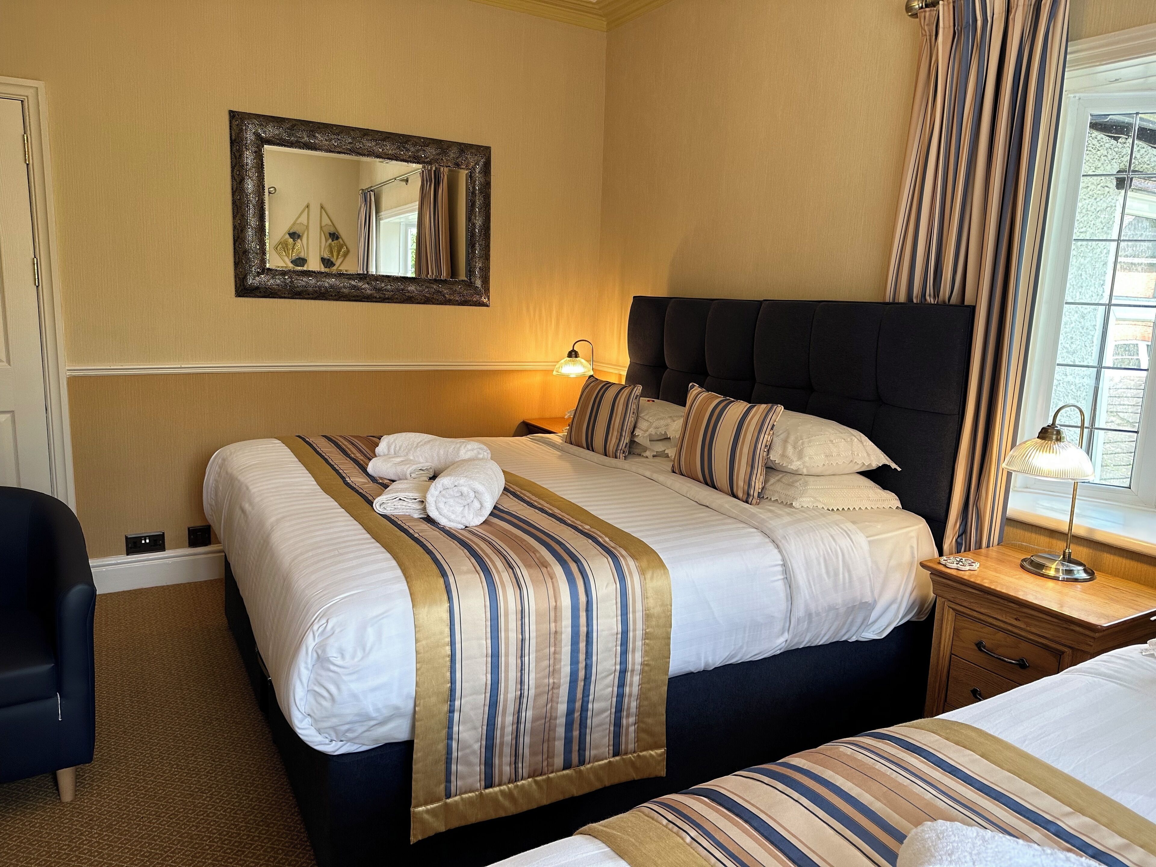 Een hotelkamer met een groot bed, een kleiner bed, een nachtkastje met lamp, een spiegel aan de muur en een raam met gordijnen.