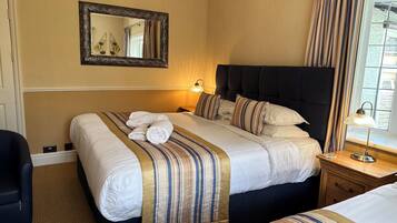 Een hotelkamer met een groot bed, een kleiner bed, een nachtkastje met lamp, een spiegel aan de muur en een raam met gordijnen.