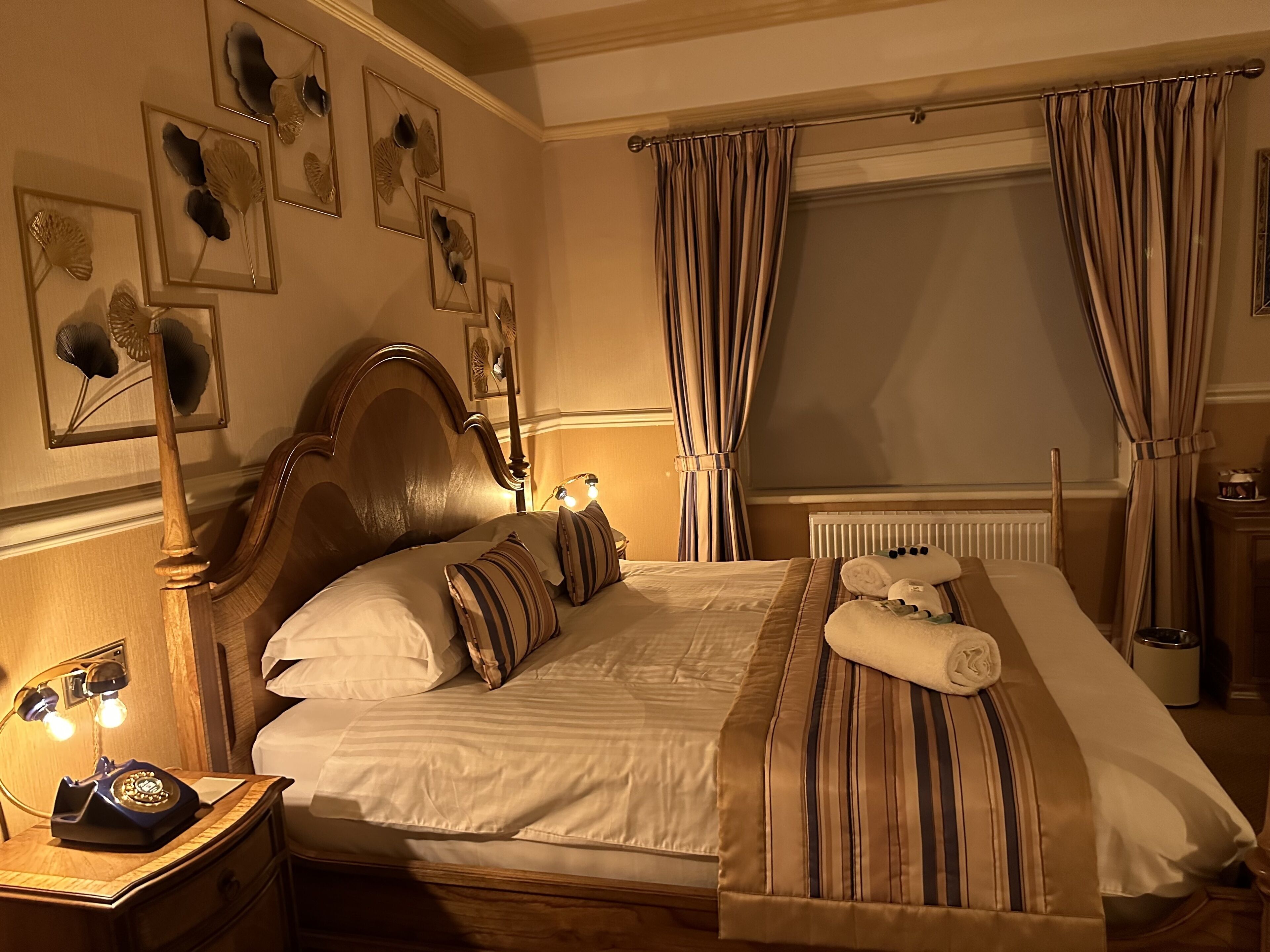 Habitación de hotel con una cama grande, cabecera de madera, mesita de noche con teléfono y ventana con cortinas.