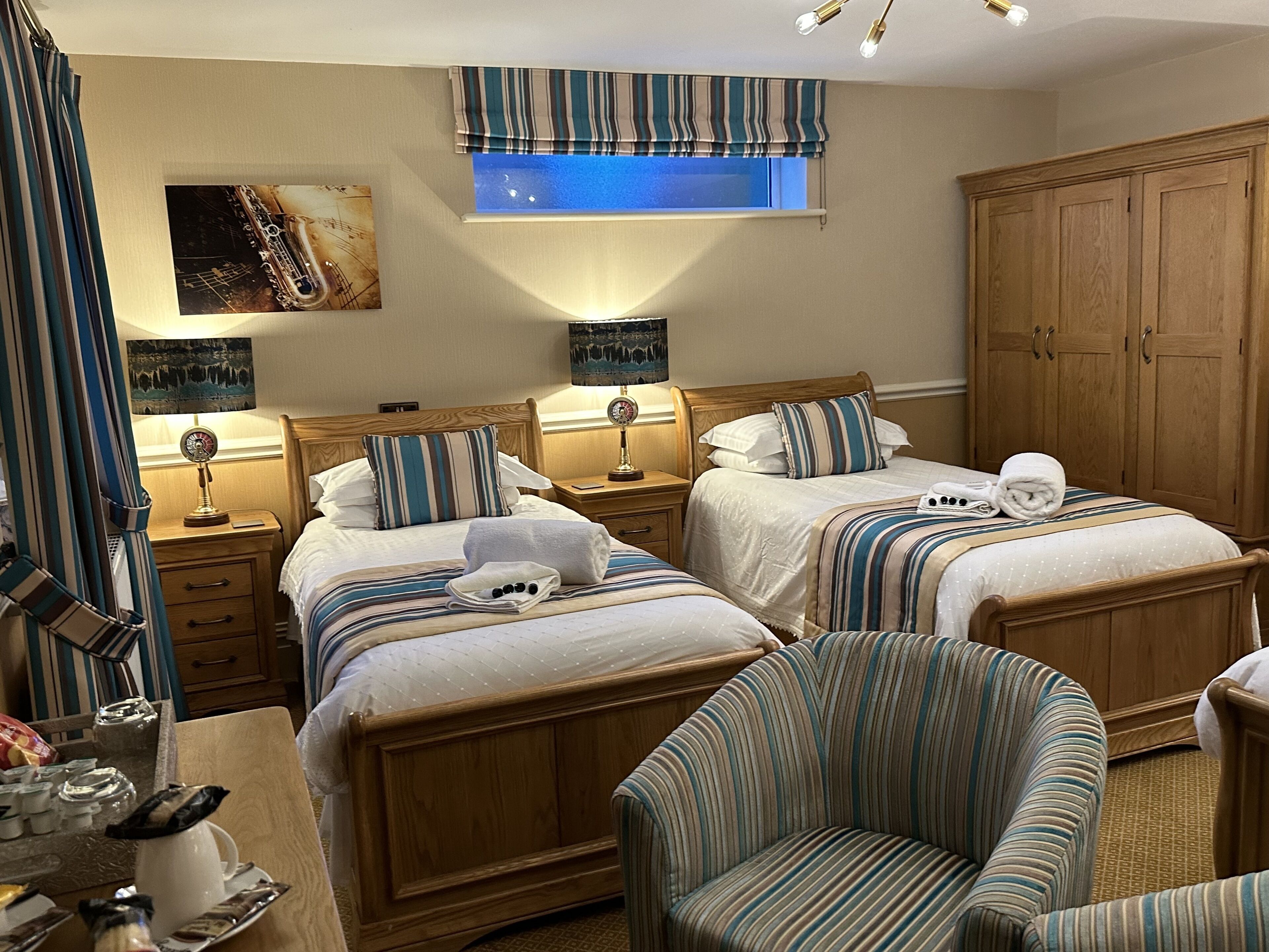 A hotel room with two beds, a wooden wardrobe, a striped armchair, and a window with a striped valance.