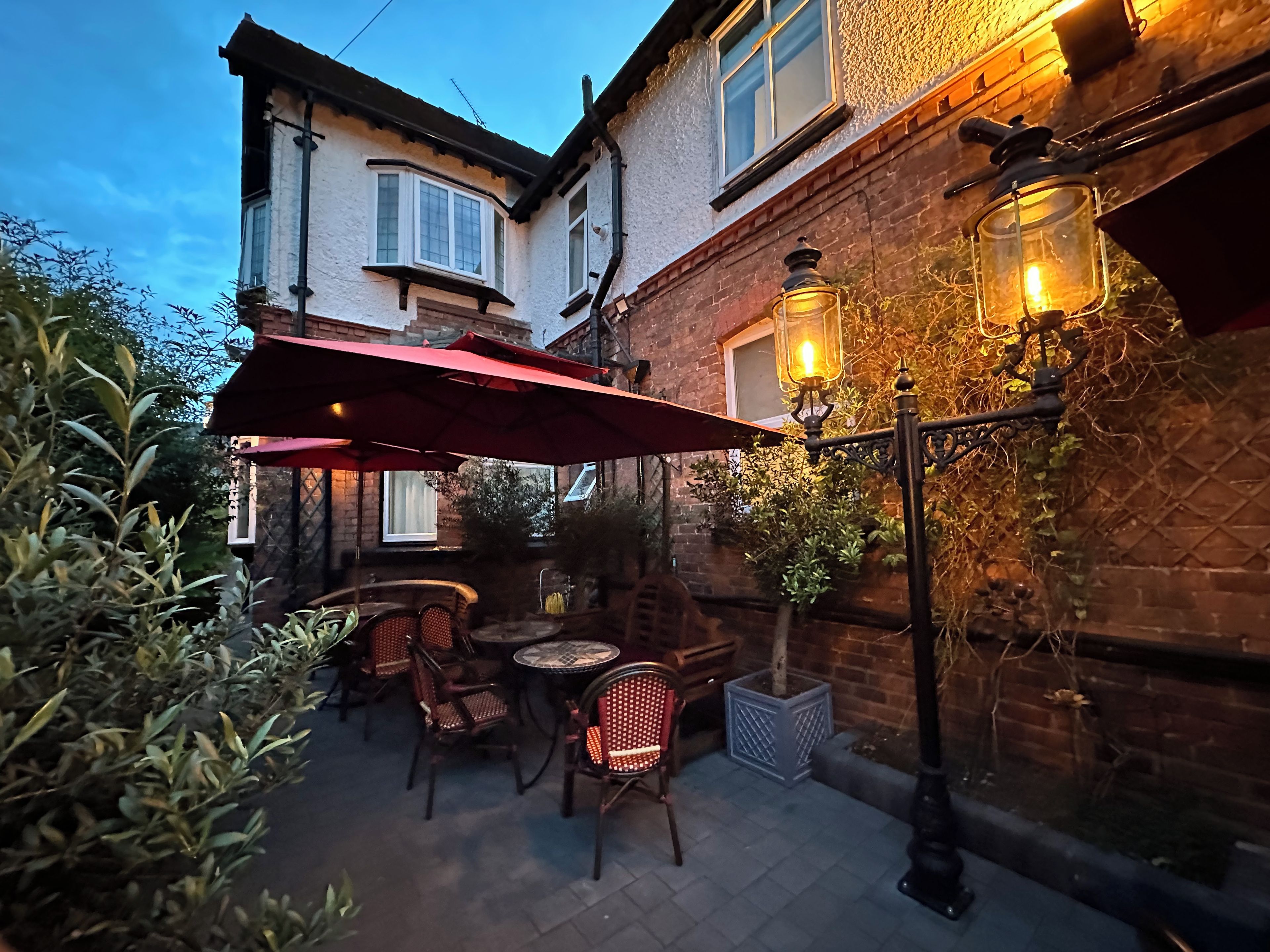 An outdoor patio area with tables and chairs, a red umbrella, and a brick building in the background.