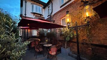 An outdoor patio area with tables and chairs, a red umbrella, and a brick building in the background.