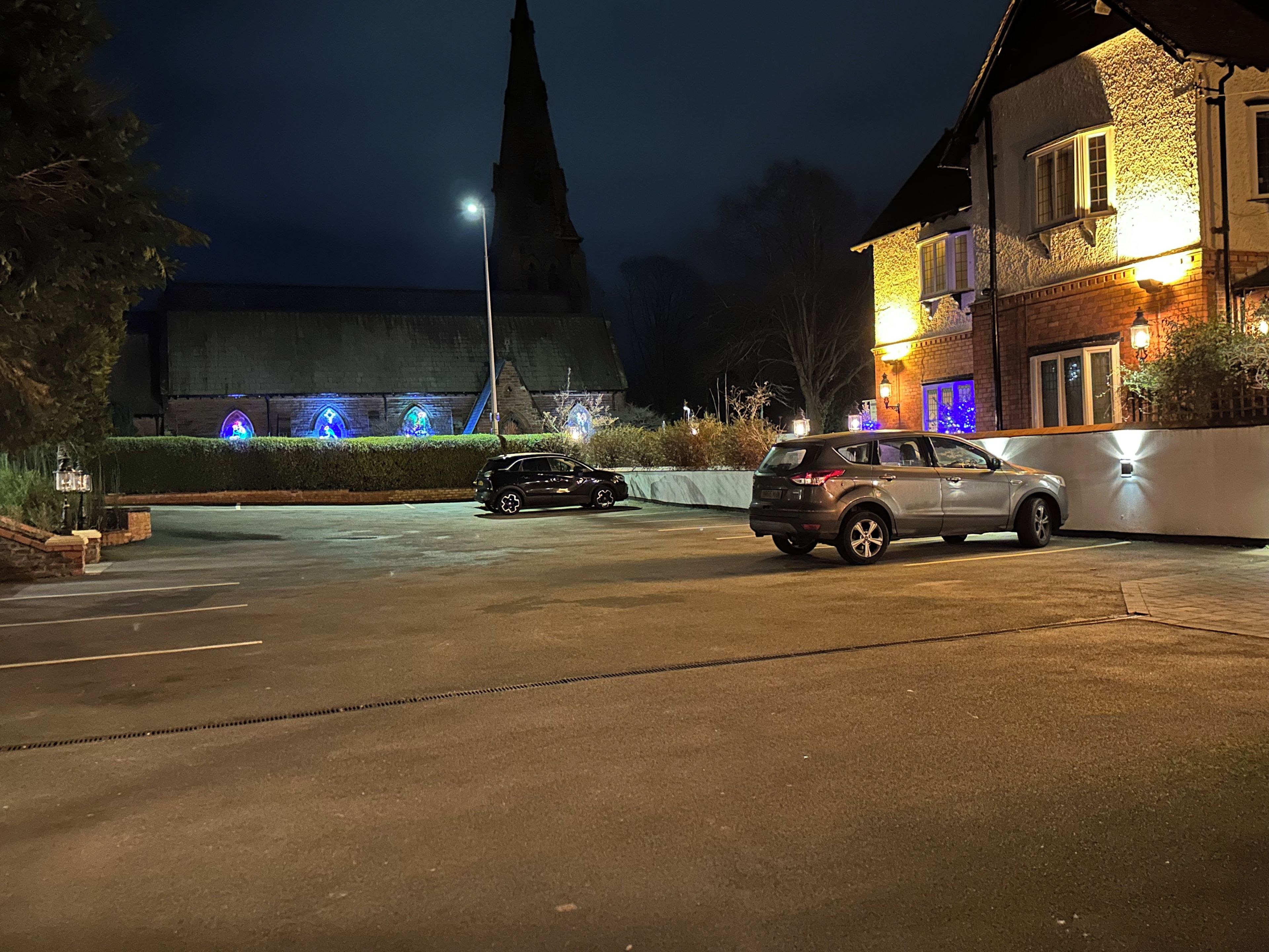 A parking lot with two cars, a building with illuminated windows, and a church spire in the background.
