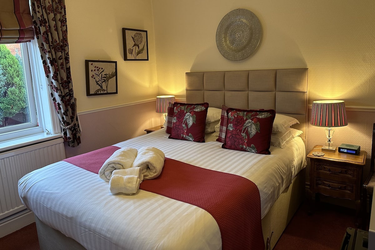 A neatly made bed with a red and white bedspread, two pillows with floral embroidery, a bedside table with a lamp, and a wall clock.
