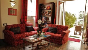 Een woonkamer met twee gestreepte banken, een salontafel met boeken en een vaas, een boekenkast met diverse objecten, en uitzicht op het groen door een glazen deur.