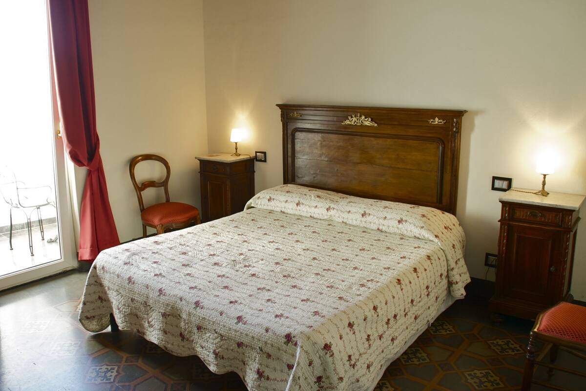 Camera da letto con testiera in legno, letto con copriletto floreale, una sedia e due comodini con lampade.