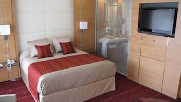 Habitación de hotel con cama, televisor, escritorio y baño visible a través de un panel de vidrio.