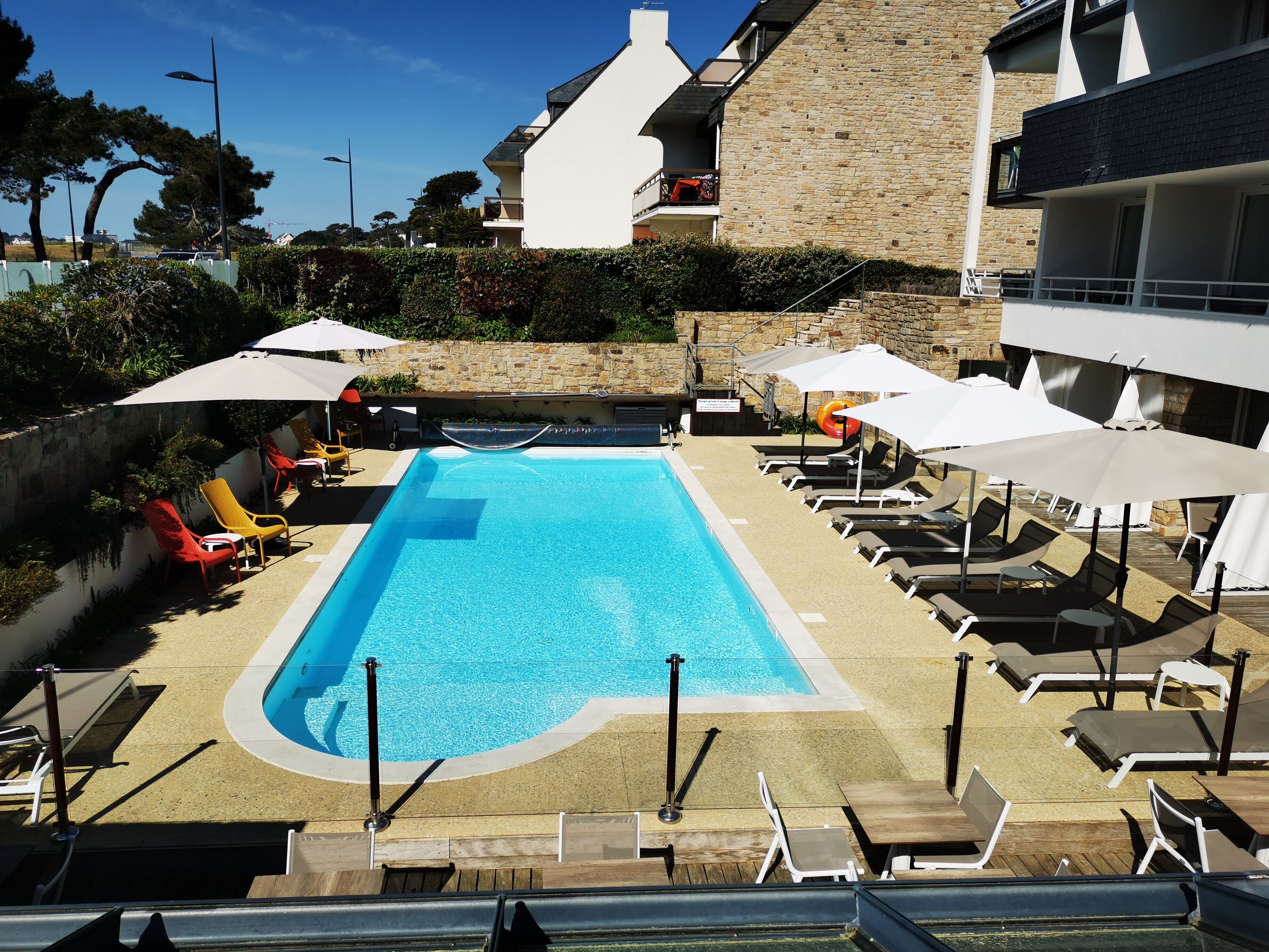 Piscina com espreguiçadeiras e guarda-sóis, cercada por edifícios de pedra e vegetação.