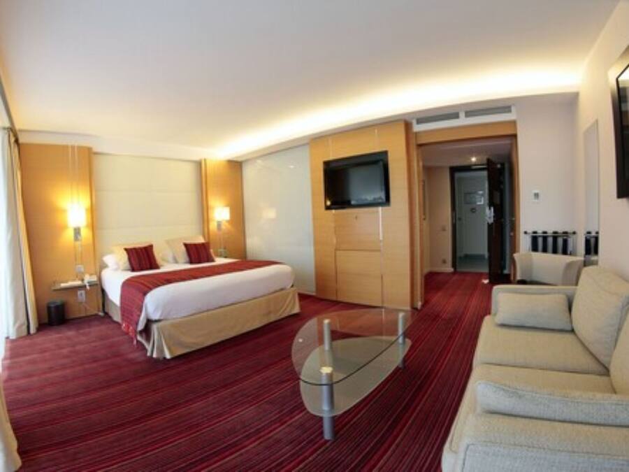 Habitación de hotel con cama, sofá, televisión montada en la pared y una mesa de centro de vidrio.