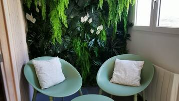Een hoekzaal met twee groene stoelen en een kleine tafel, een groene wand met hangplanten, en een raam met natuurlijk licht.