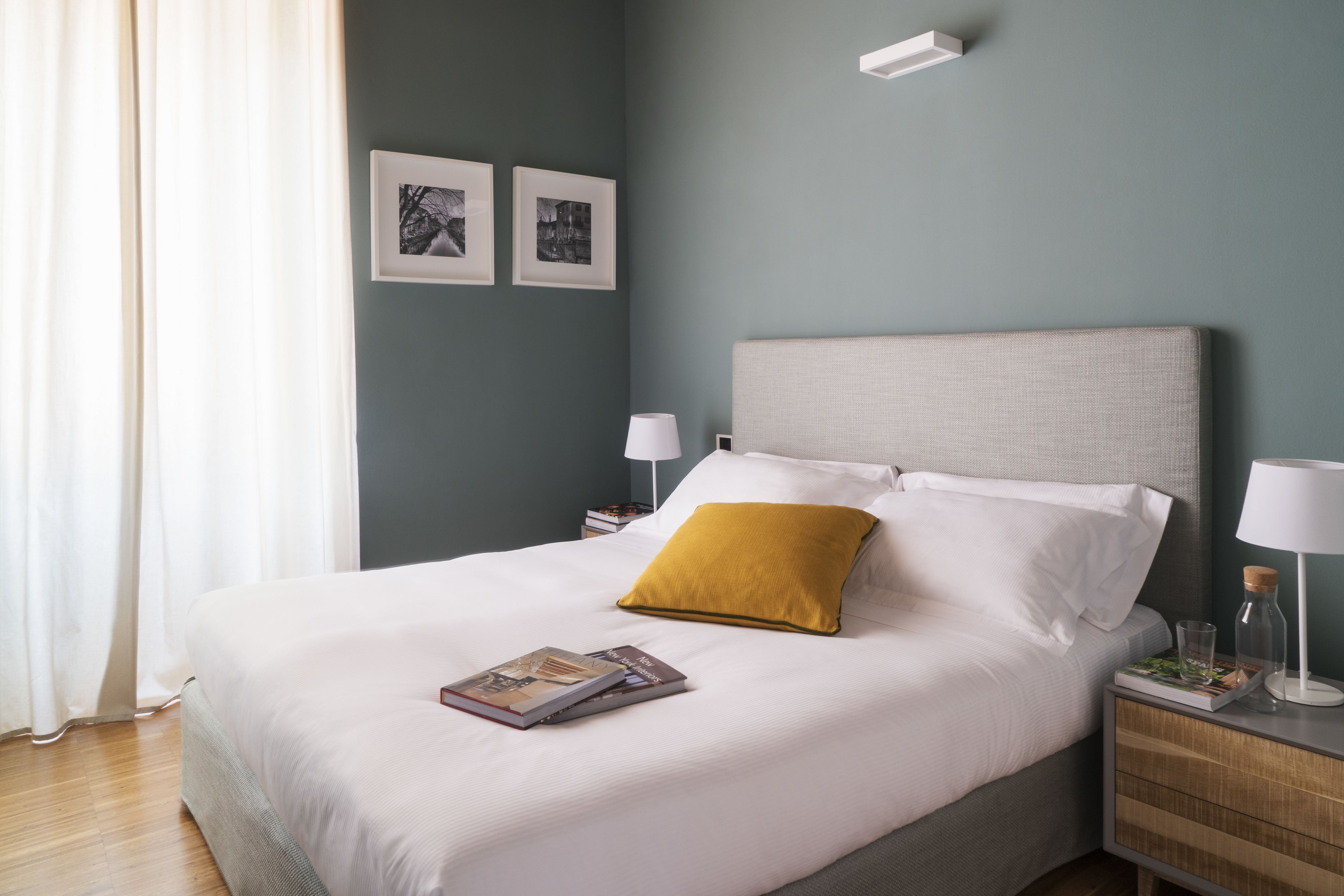 A neatly made bed with white linens and a mustard-colored pillow, a bedside table with a lamp, and two framed pictures on the wall.