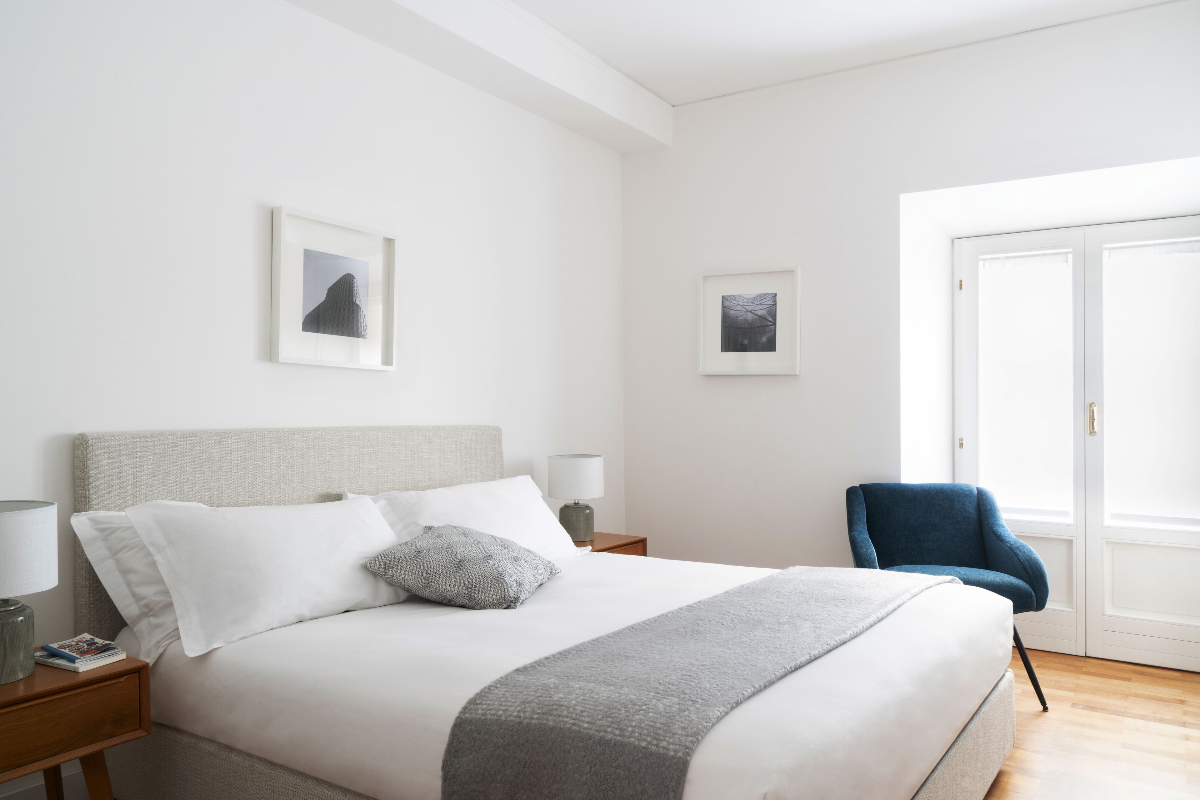 A neatly made bed with white and gray linens, a gray throw blanket, and two pillows. There is a blue chair to the right and a small table with a lamp and a book beside the bed.