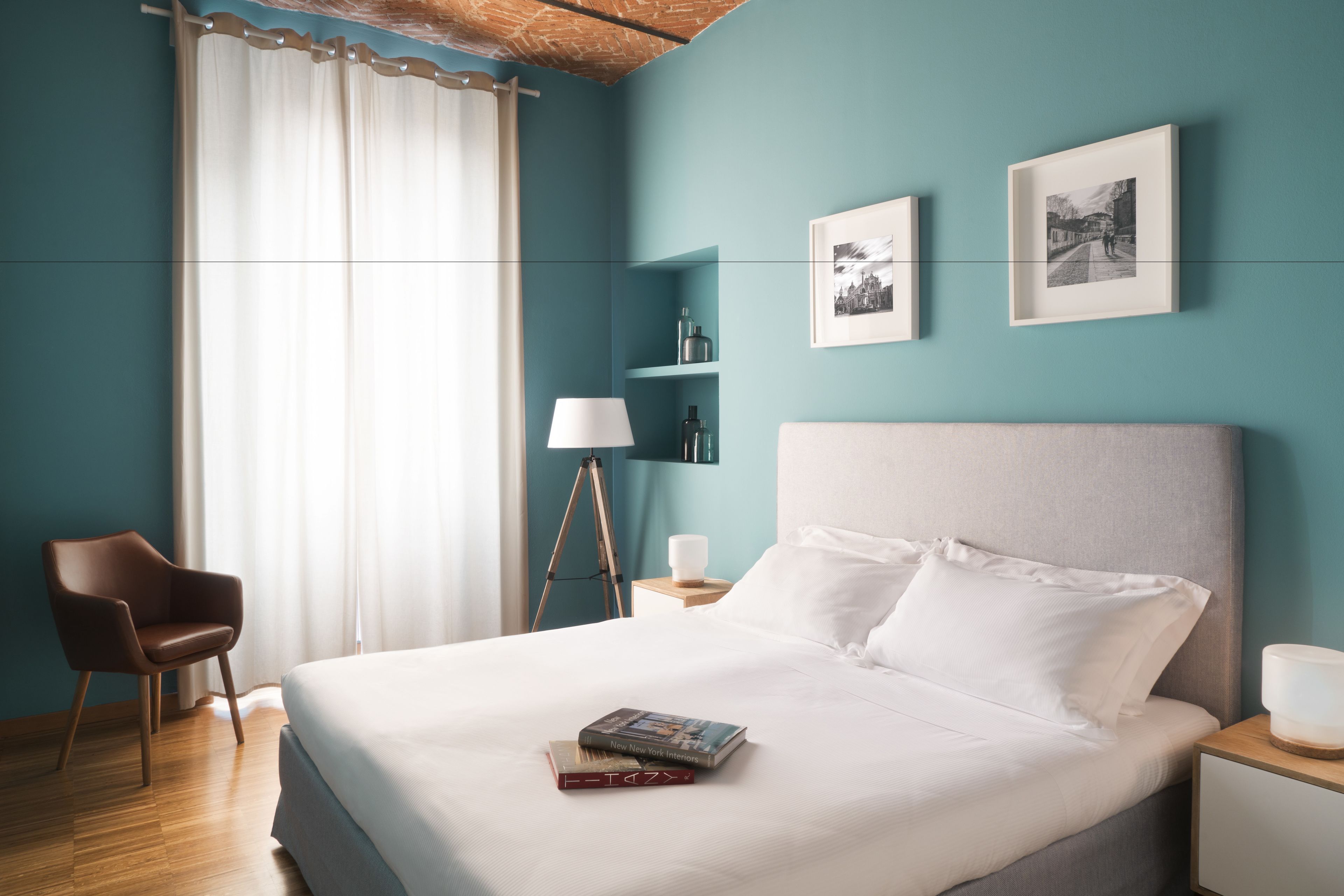 Una cama bien hecha con almohadas y un libro, una silla, una lámpara y cuadros en paredes color turquesa.