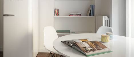 Una cocina moderna con armarios blancos, horno empotrado y una mesa de comedor blanca con sillas. Hay libros y objetos decorativos en los estantes sobre la mesa.